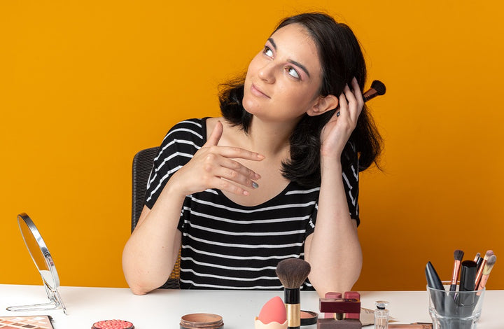 Women looking up and holding makeup brush and makeup products kept around her | dusky skin complexion